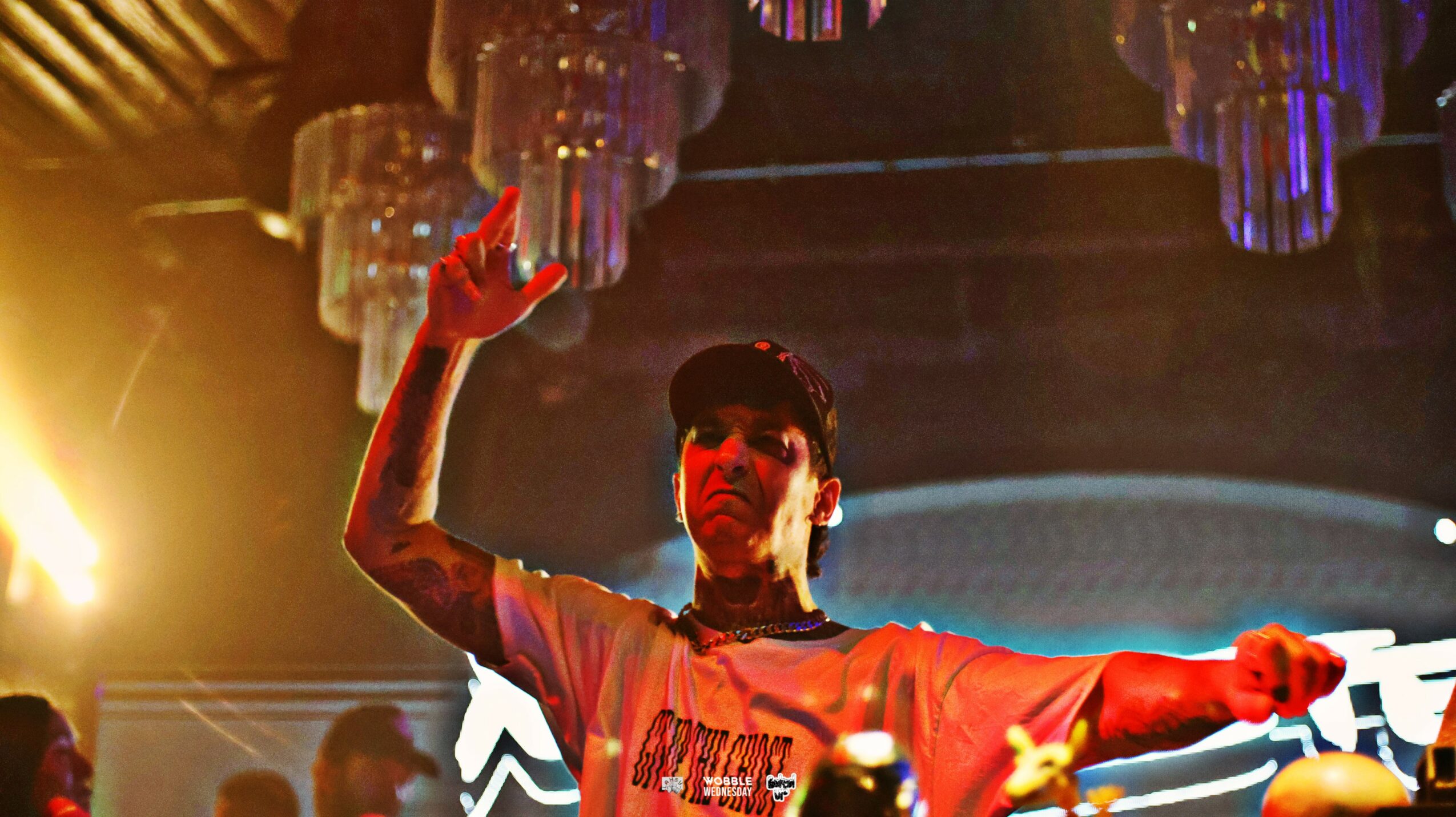 Person with tattooed arm raising hand in a lively indoor setting with colorful hanging decorations and lighting