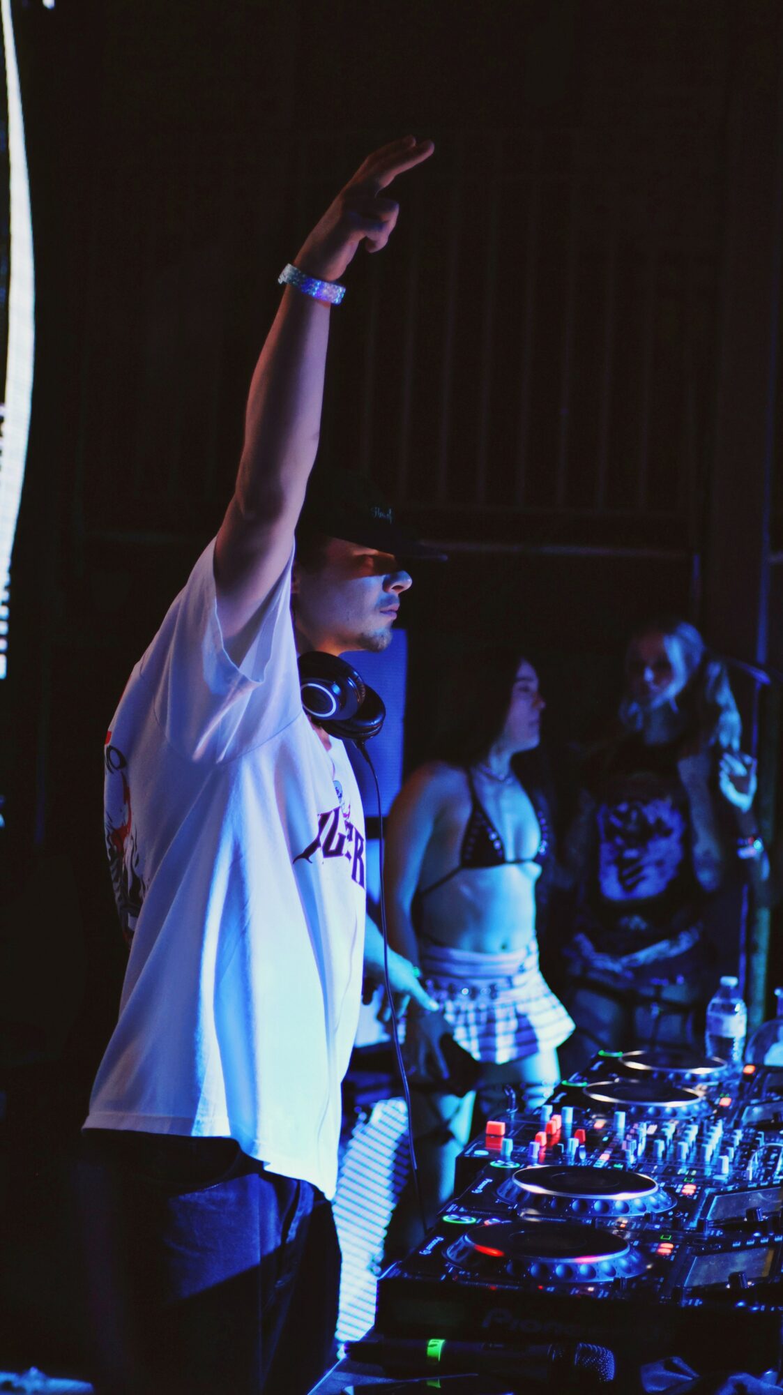 Person with raised arm wearing a white shirt and glasses at a DJ booth with people in background.