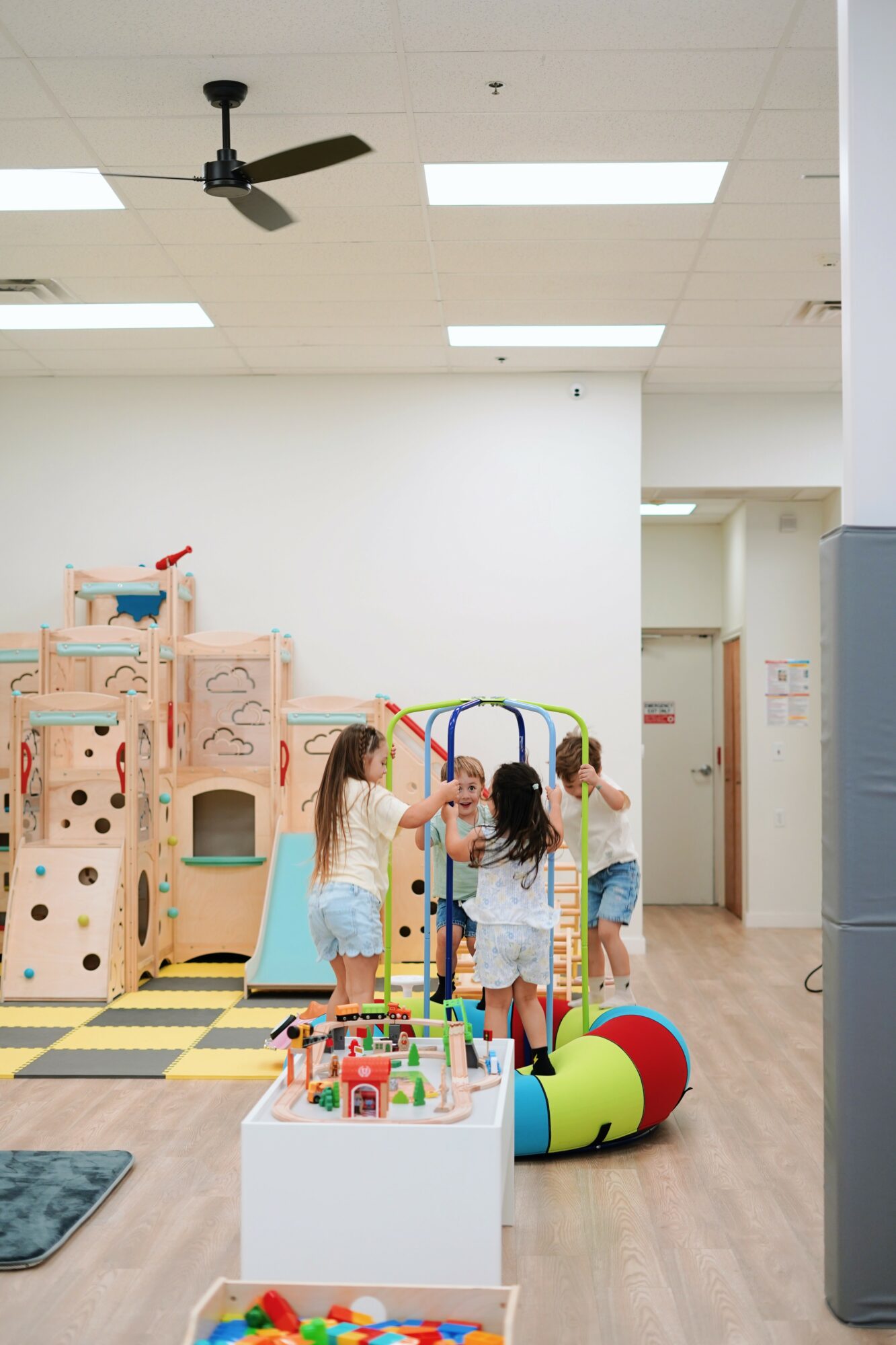 Children playing with toys in an indoor play area with a wooden structure, colorful balls, and a white table, ceiling fan visible.