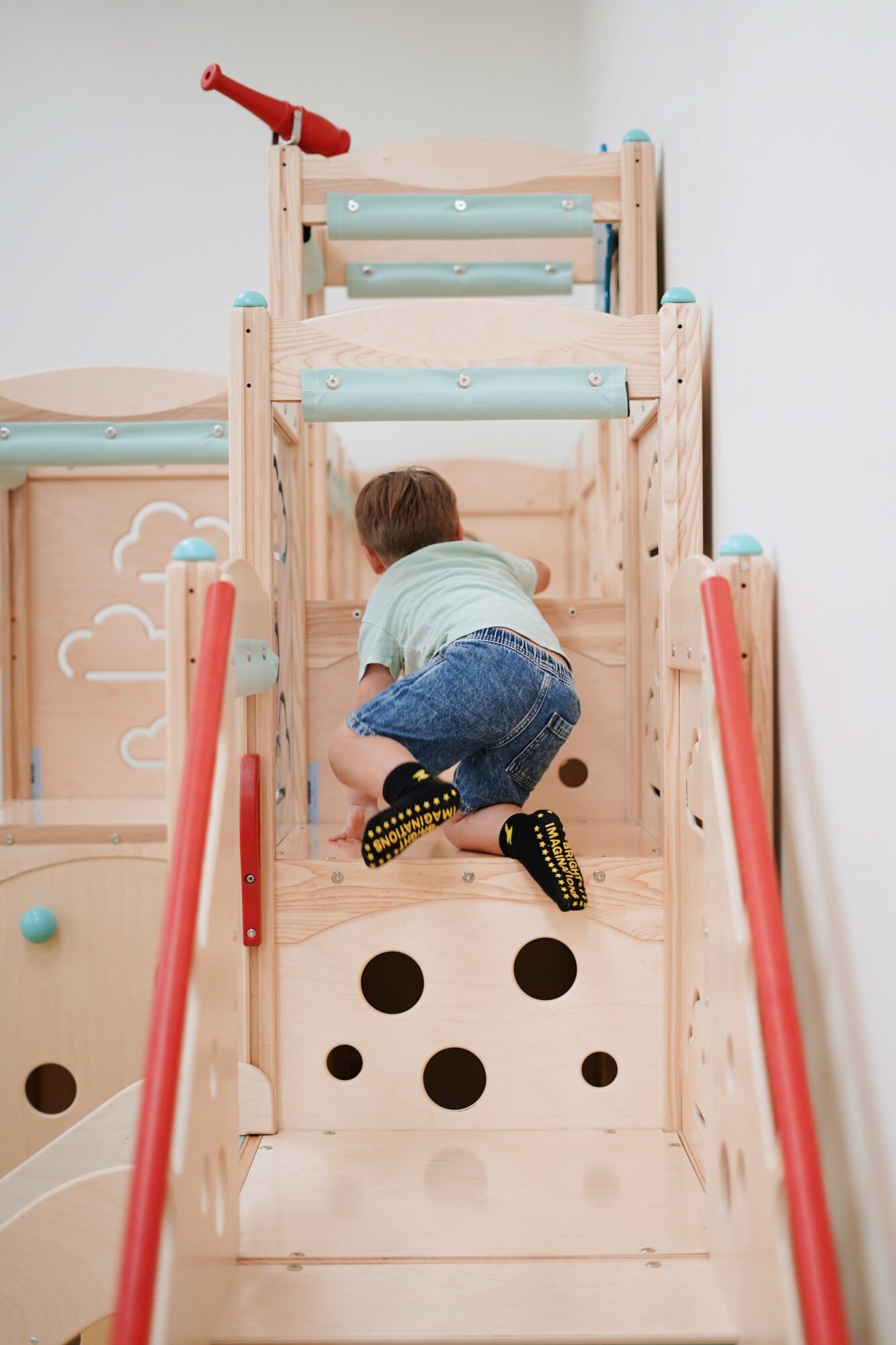 Child climbing a wooden indoor play structure with red and blue accents, facing away from the camera.