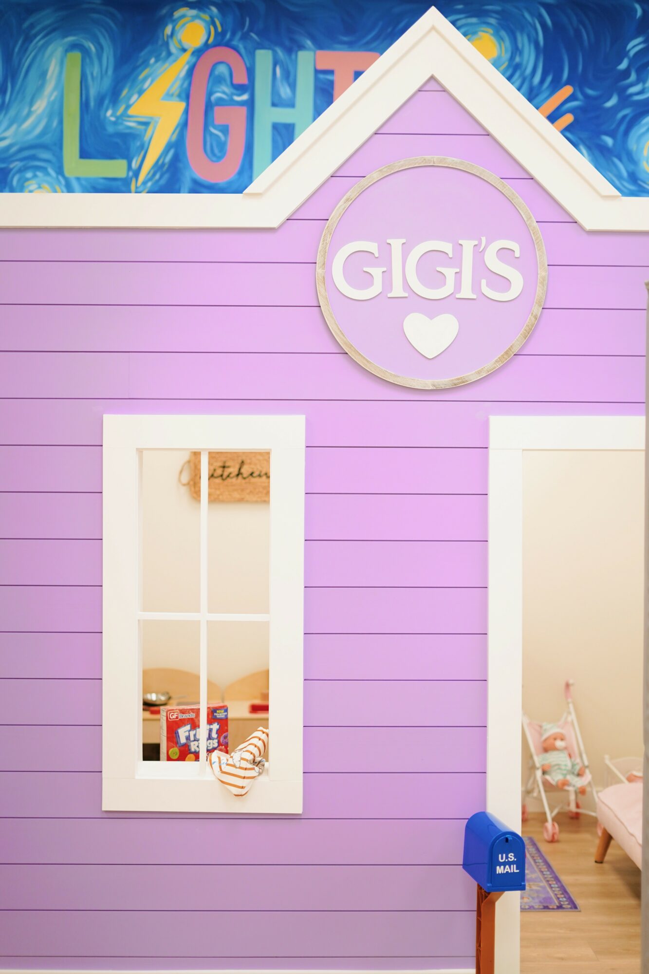 Pink house with a circular sign reading 'GIGIS' and a heart, window showing toys inside, and a blue mailbox outside.