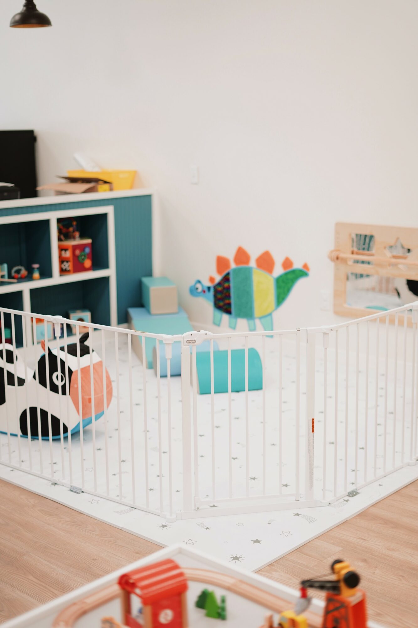Children's play area with colorful animal and block toys, white safety gate, and storage shelves, against a white wall.