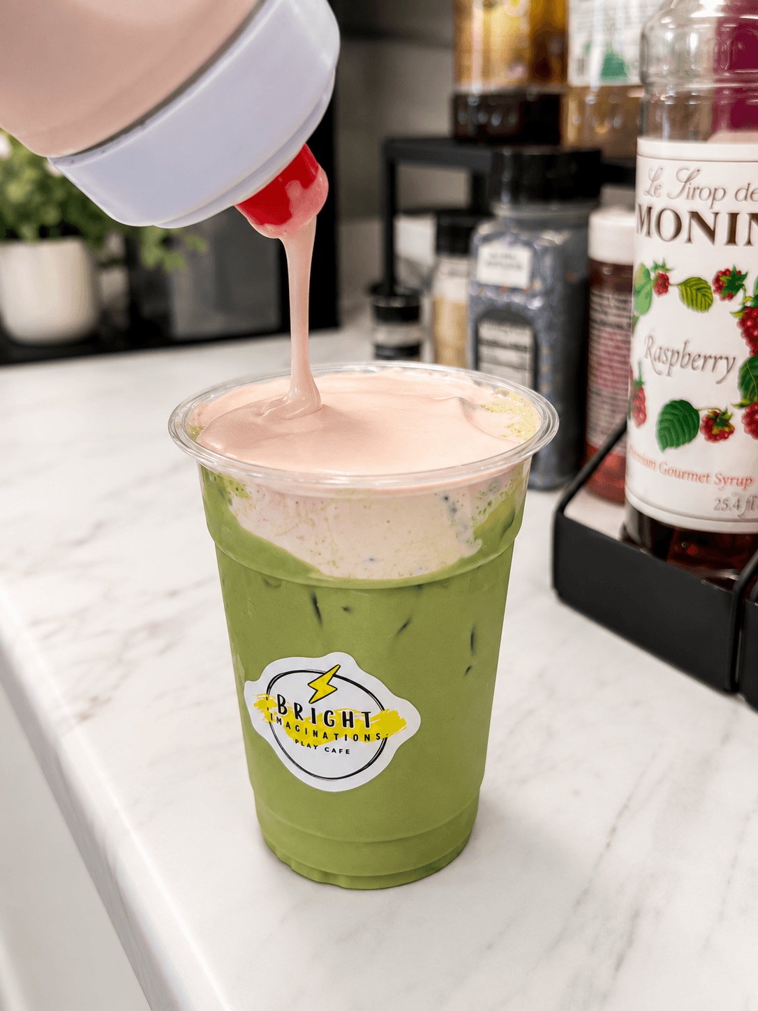 Green beverage in a clear cup with pink foam being poured on top, on a white surface with bottles in background.