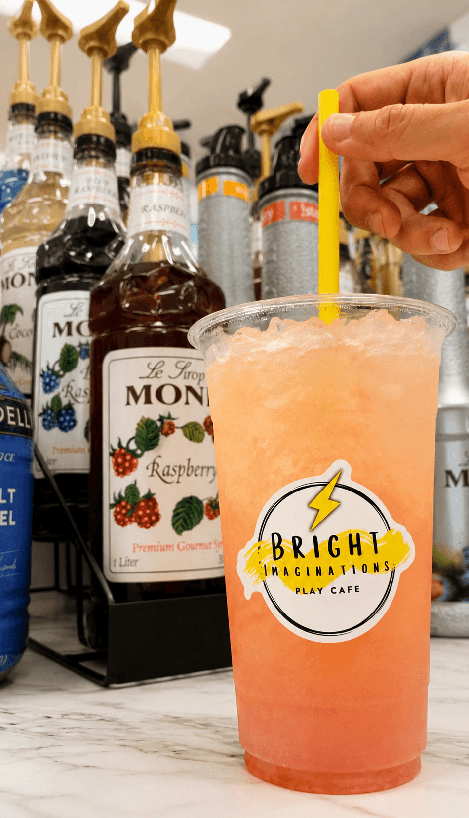 Hand holding a yellow straw in a pinkish-orange drink with a Bright packaging sticker, bottles in the background