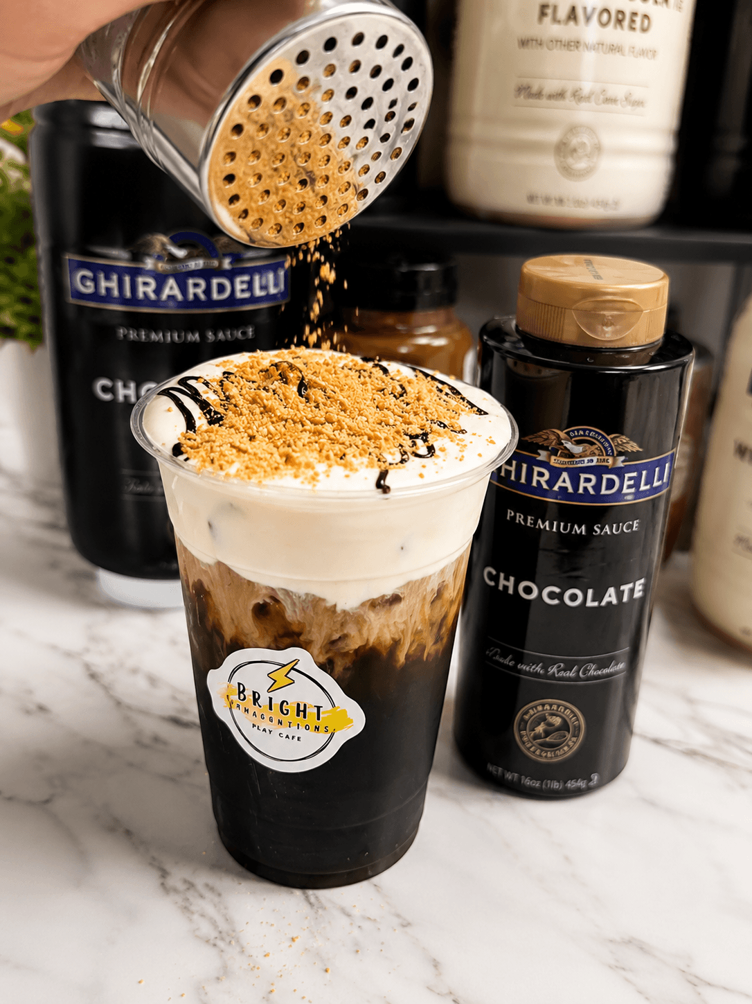 Person sprinkling crushed cookies or toppings onto a layered chocolate dessert in a glass, with chocolate syrup and sauce bottles in background.