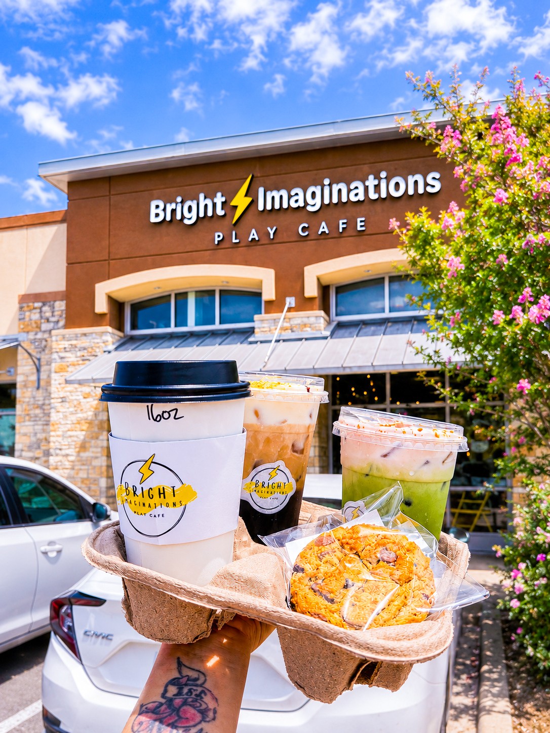 Bright Imagination Play Cafe building with a sign, blue sky, pink flowering tree, and outdoor seating area.