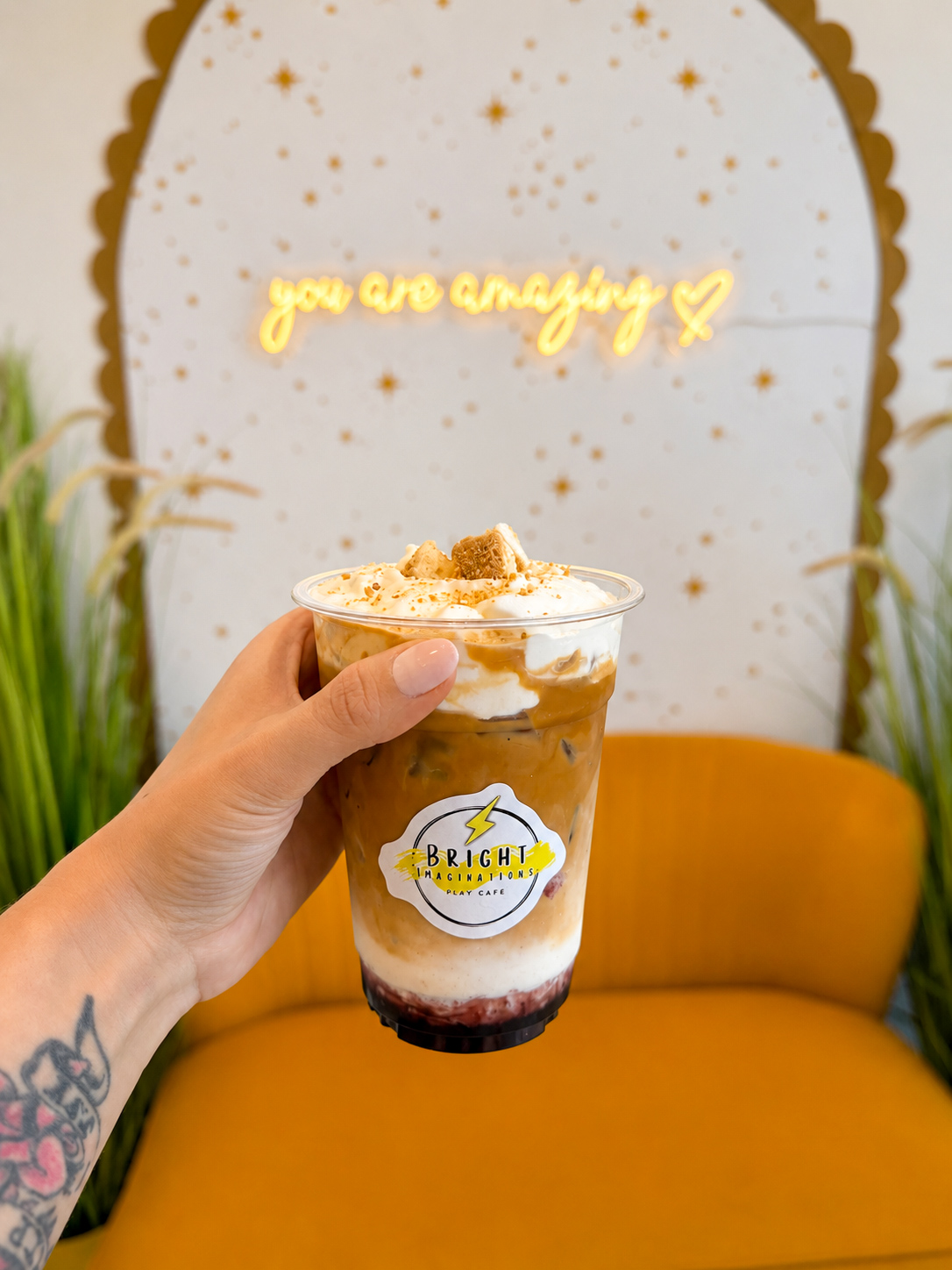 Hand holding a layered iced coffee with whipped cream and caramel, in front of a decorated wall with a yellow neon sign, and a yellow couch.