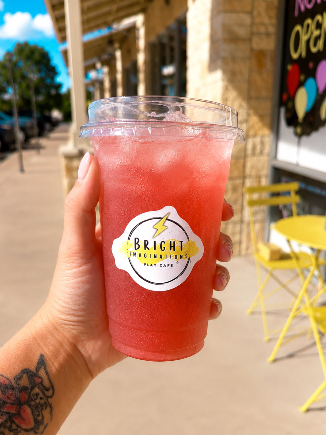 Hand holding a pink frozen drink in a clear cup with a Brich logo, outdoor cafe setting with yellow chairs.
