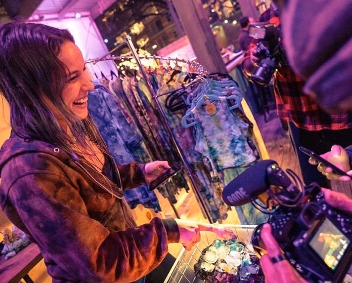 Woman smiling at a display of colorful clothing and accessories in a store window at night.