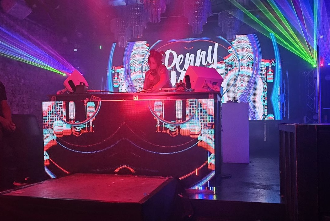 DJ performing on stage with colorful lights and a digital backdrop displaying the word 'Denni'.