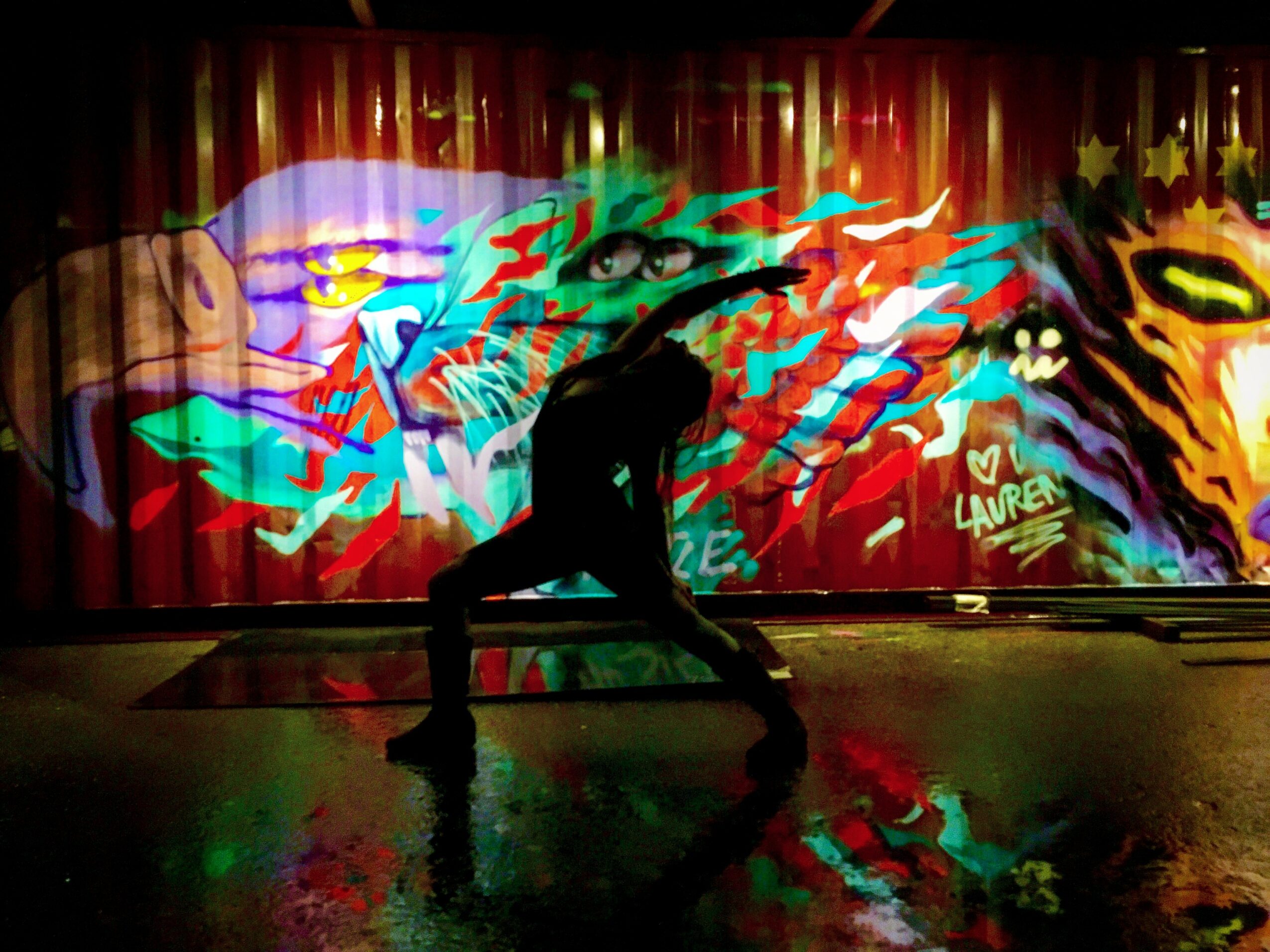 Person performing on stage with colorful graffiti-style backdrop, silhouette visible, stage reflected on floor, vibrant and dynamic scene.