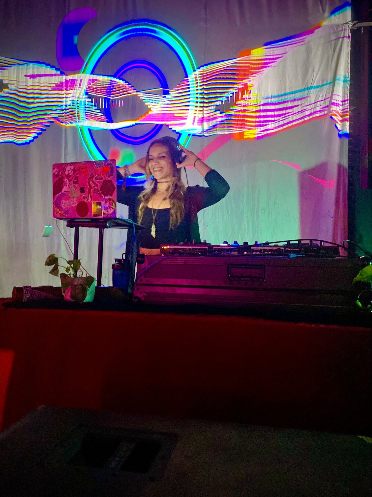 Person with long hair behind DJ equipment, colorful neon lights and abstract patterns in background, plant on table, dark setting.