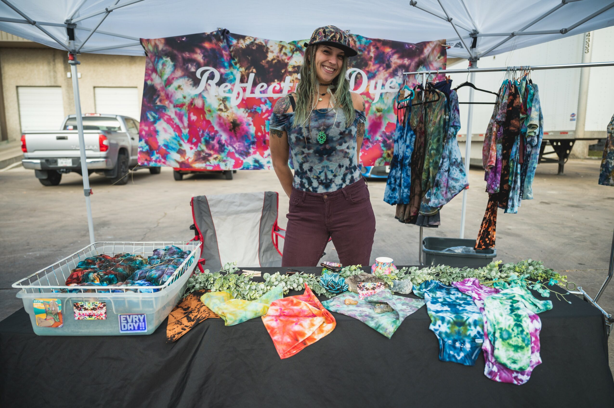 Woman standing behind a table with colorful fabric and clothing at an outdoor market stall, with a banner in the background.