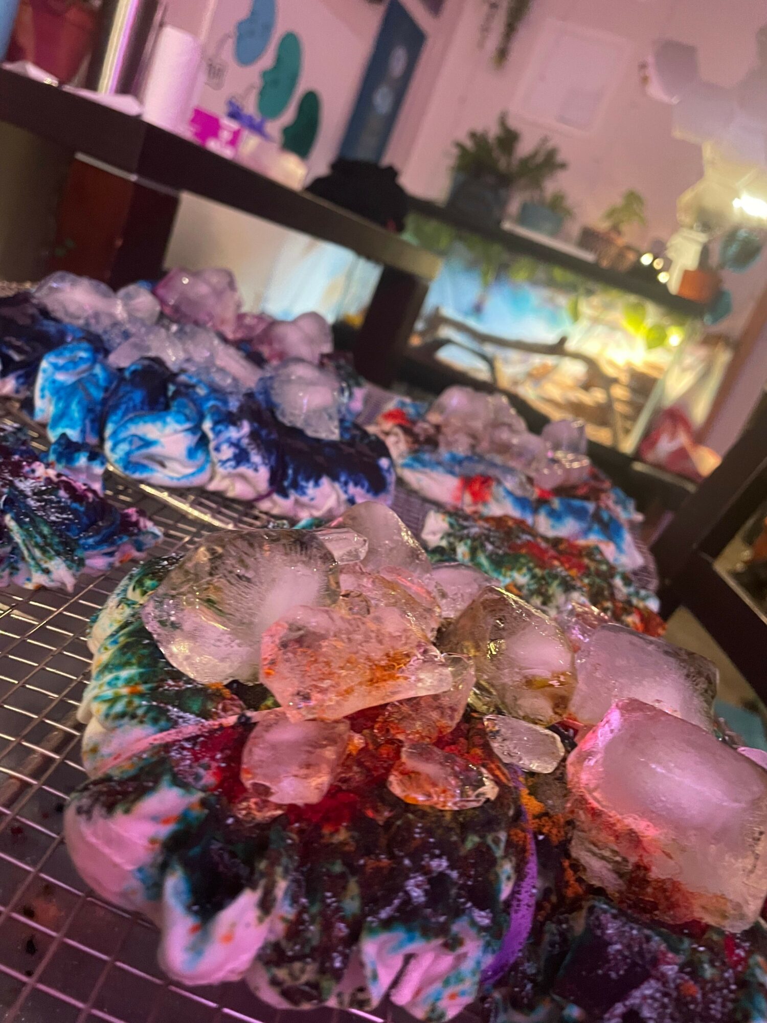 Ice cubes and colorful crushed ice on a wire rack, with a blurred background of a window and interior.