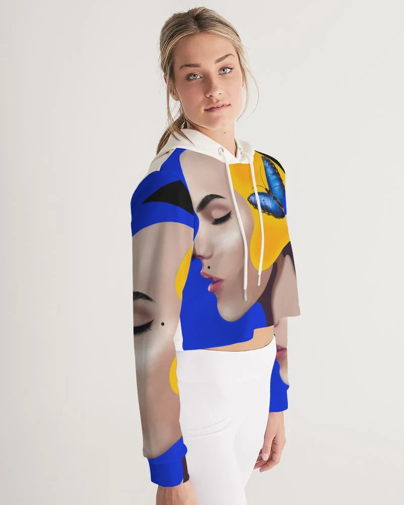 Young woman with blonde hair wearing a colorful abstract patterned top and white pants, standing against a plain background.