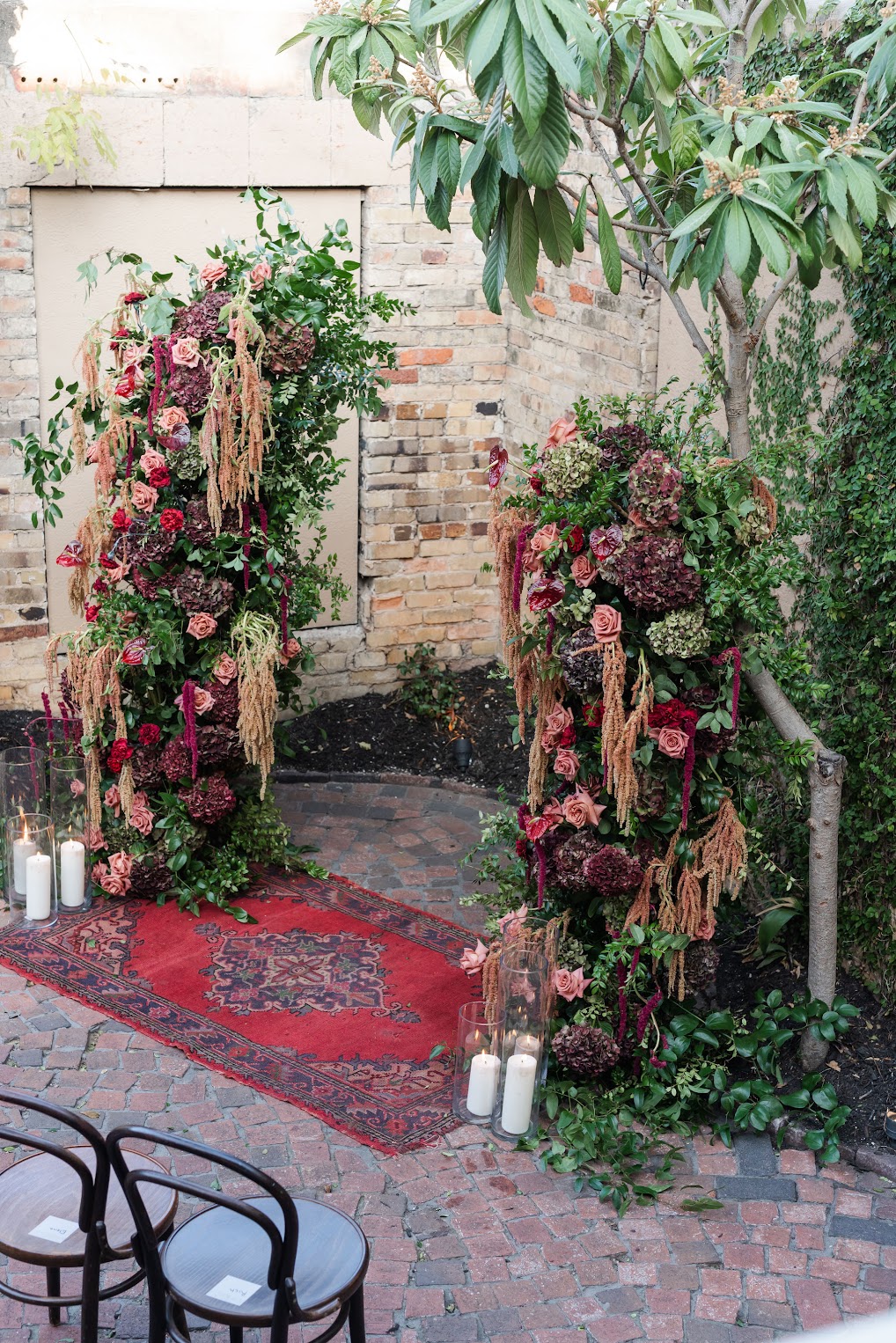 Decorative floral arch with pink and purple flowers, candles, and a red rug on cobblestone ground against a brick wall.