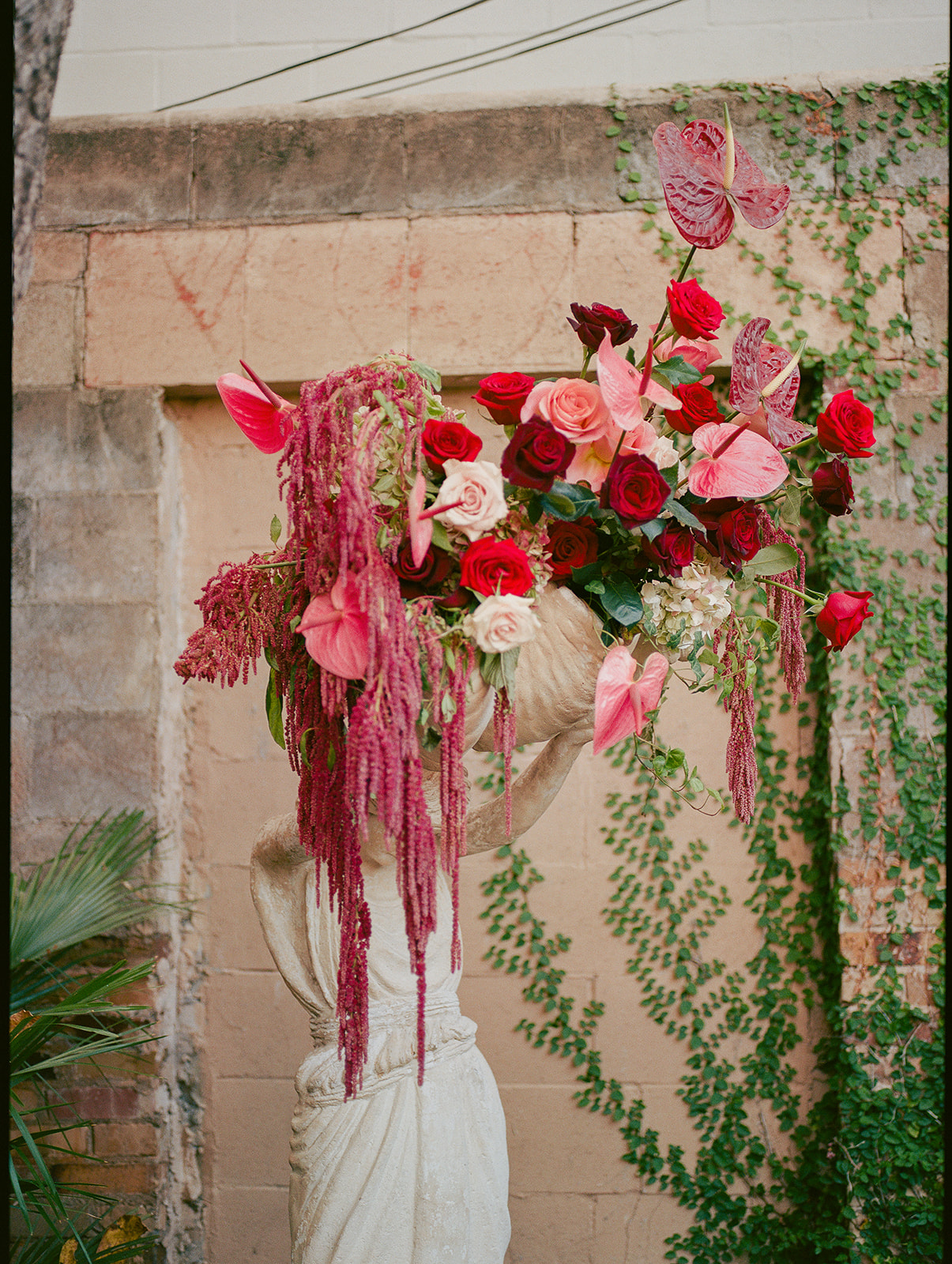 Vase with pink and red roses and greenery against a brick wall, with hanging pink flowers and vines nearby.