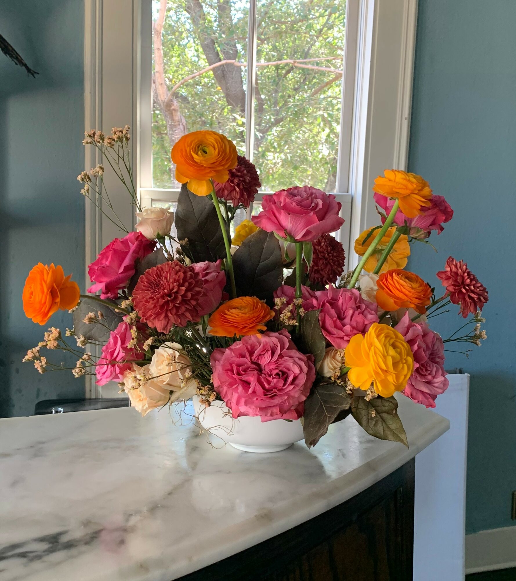 Colorful flower arrangement with pink, orange, and yellow flowers in a white vase on a marble surface, window in background.