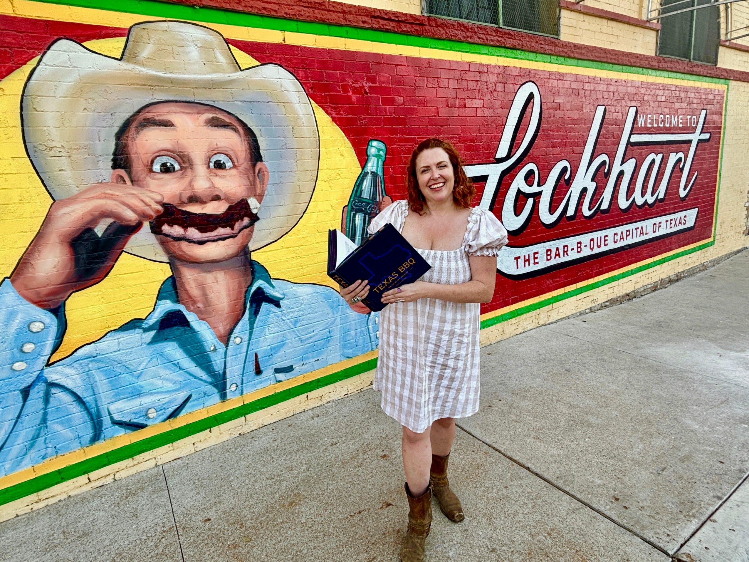 Woman in a white dress holding a book, standing in front of a colorful mural with a man in a cowboy hat, smiling.