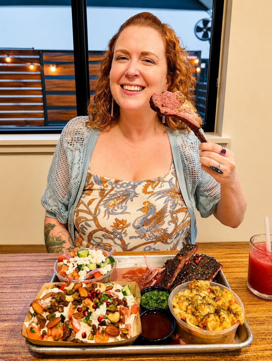 Woman with curly red hair smiling, holding a piece of meat on a fork, seated at a table with food, in front of a window.