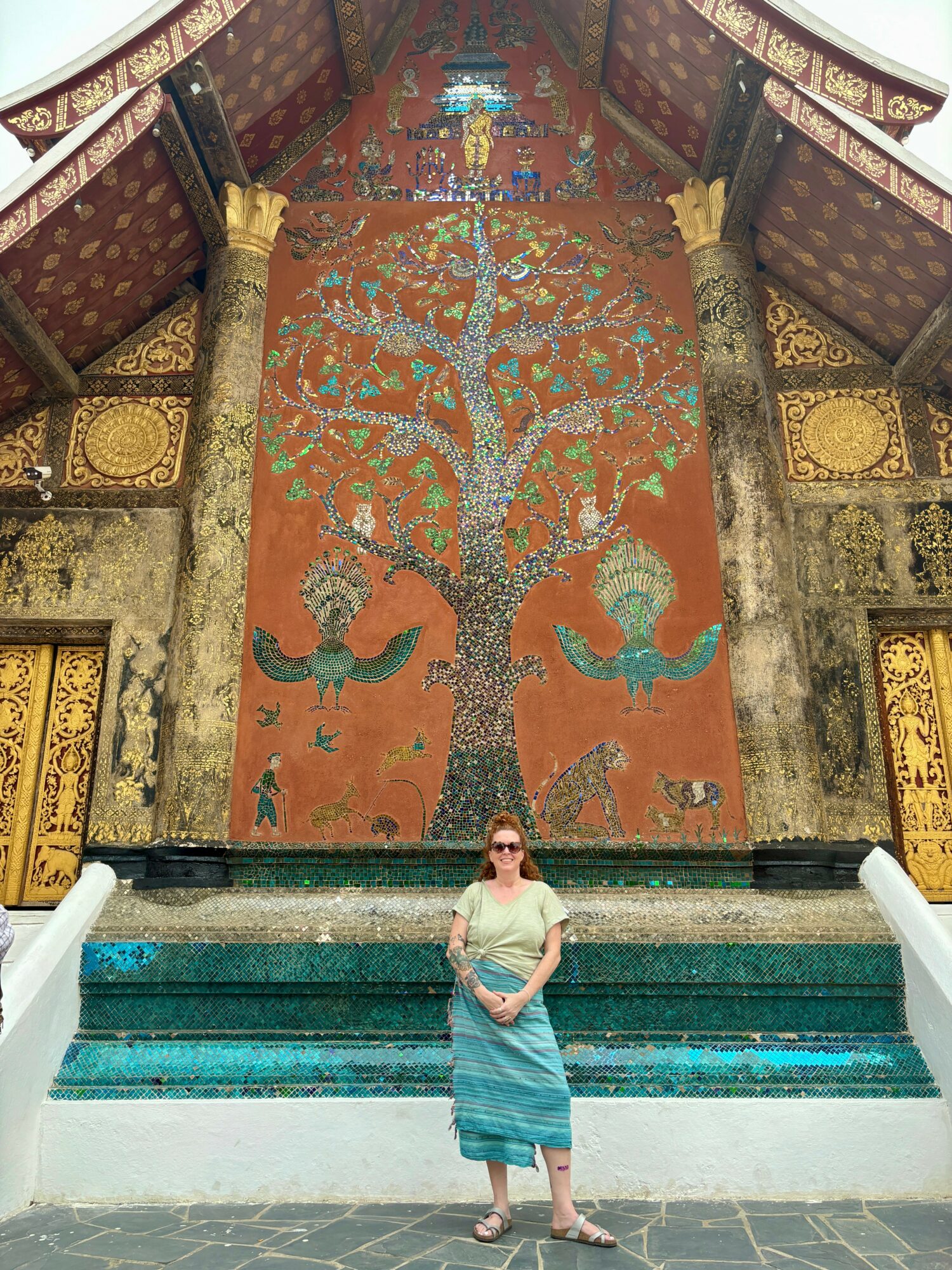 Woman in sunglasses and a striped skirt stands in front of a large decorative wall hanging with a tree design, at a temple.