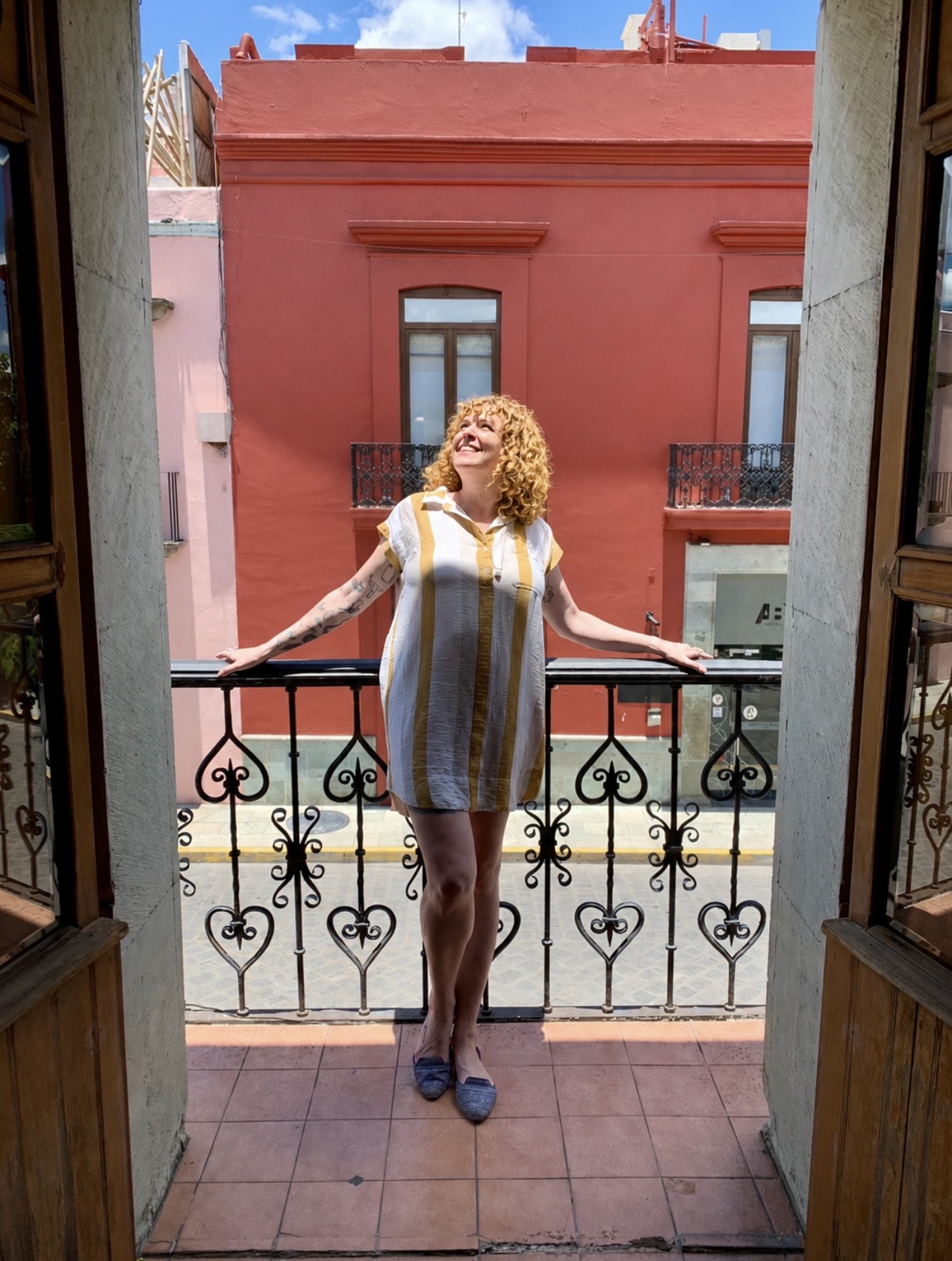 Woman with curly hair standing on balcony, arms outstretched, smiling, pink building in background.