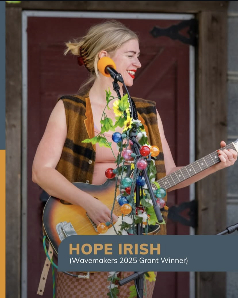Woman with blonde hair playing guitar, smiling, with colorful decorations and a microphone, outdoors, wooden background.