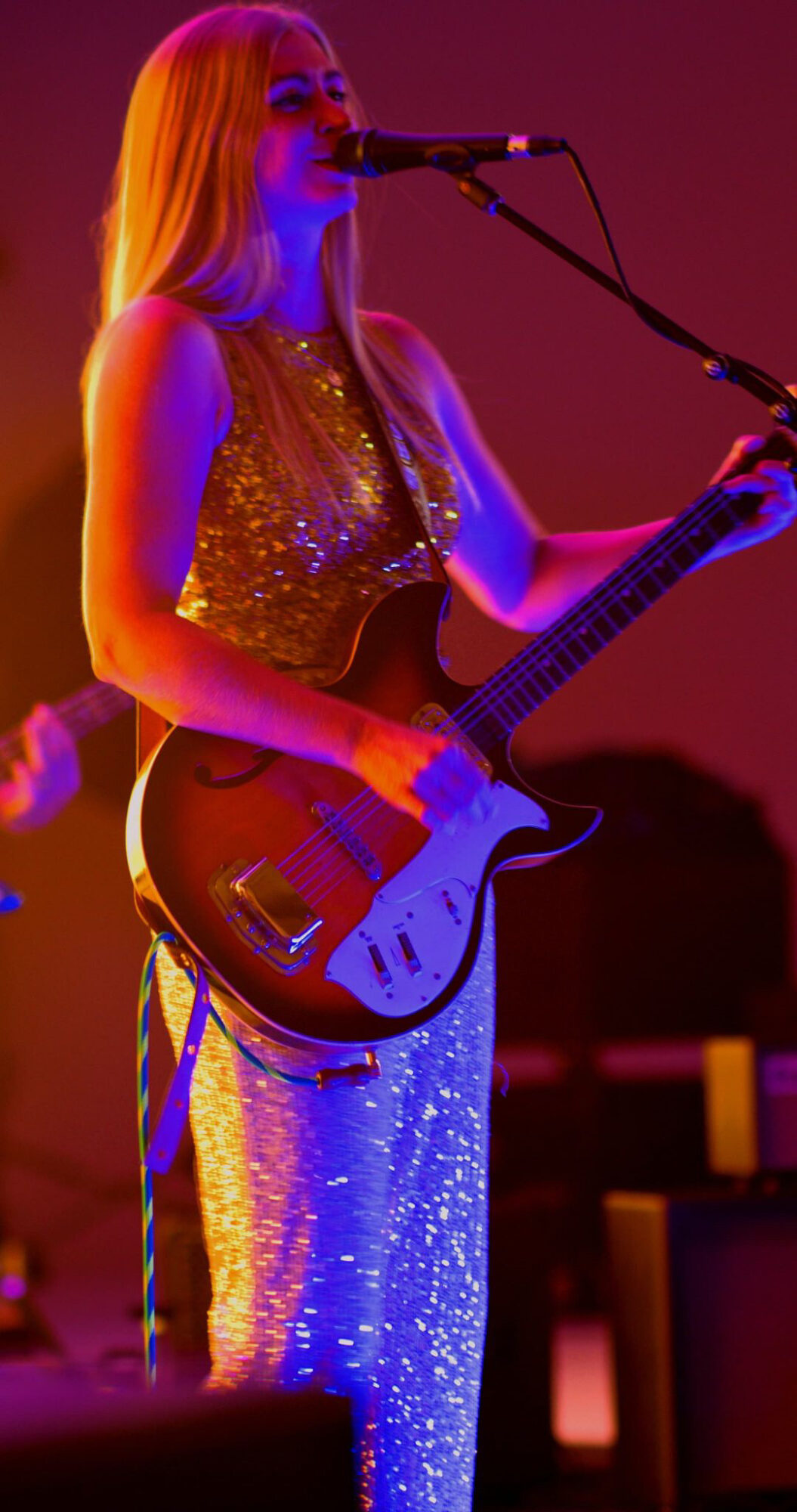 Female musician singing into microphone and playing guitar on stage, wearing a sparkly dress, with colorful stage lighting.