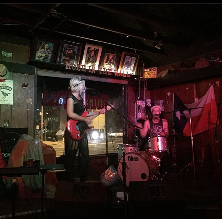 Two musicians perform on a dimly lit stage with framed photos and signs behind them, one playing guitar and the other on drums.