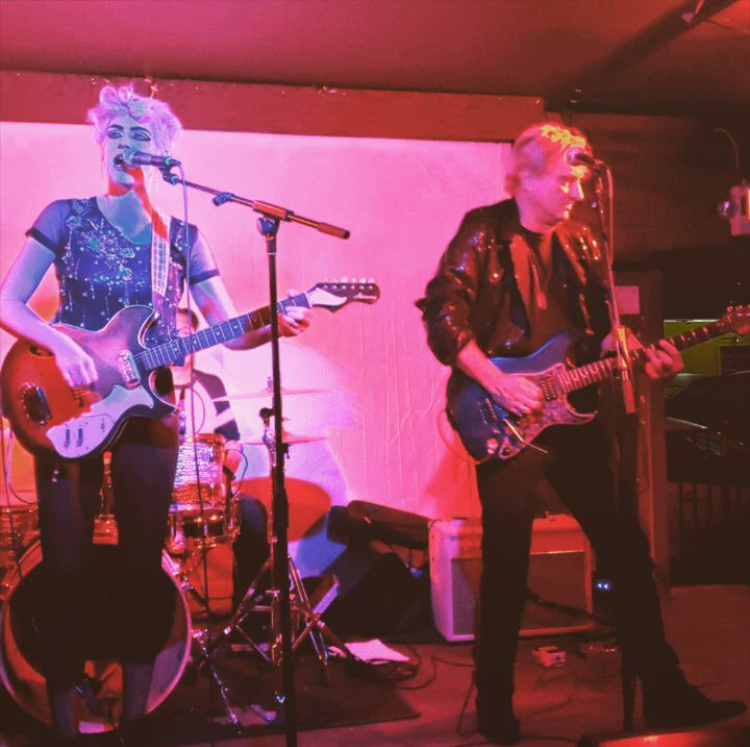 Two musicians perform on stage with guitars, a drum set, and microphones, in a dimly lit venue with a pink background.