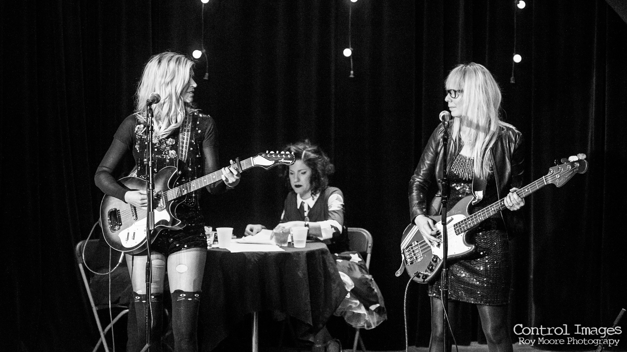 Two women playing guitars on stage with a girl sitting at a table between them, in a black-and-white setting.