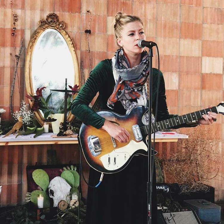 Young woman with blonde hair in a bun singing into a microphone while playing an electric guitar indoors, with a mirror and plants behind her.