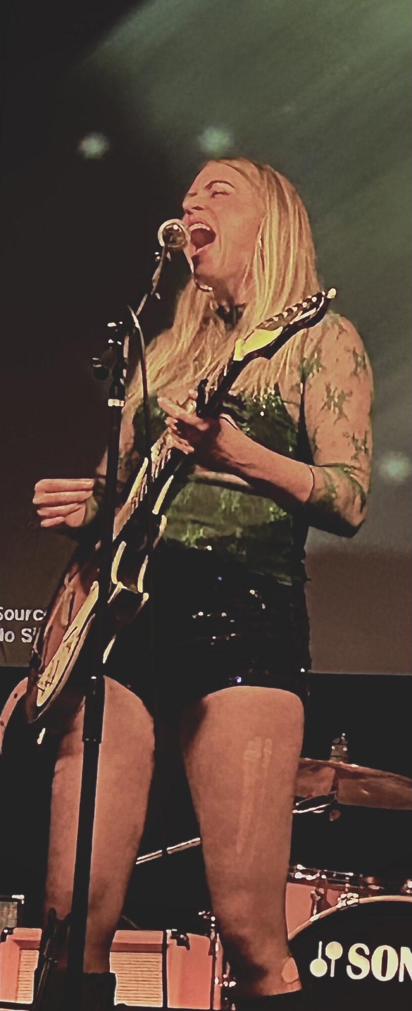 Woman singing and playing guitar on stage, wearing a floral top and shorts, with a microphone and drum set in background.