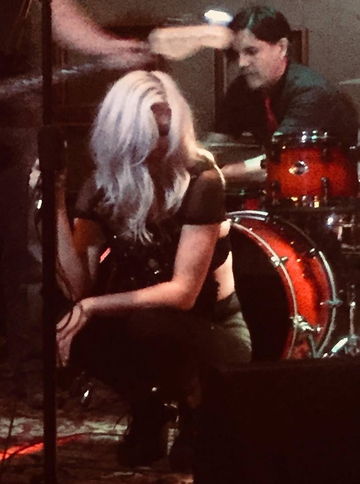Person with long, wavy blonde hair sitting near a drum set, with a man in the background looking at her.