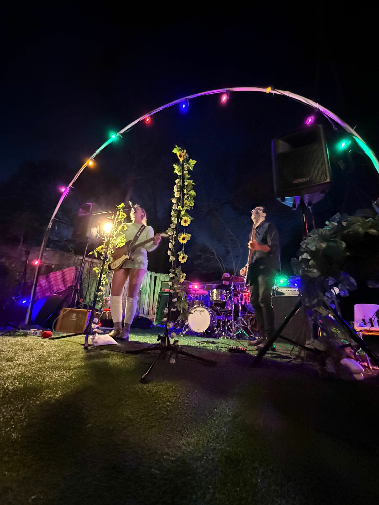 People performing on an outdoor stage at night with colorful lights and a rainbow arch, surrounded by musical instruments and equipment.