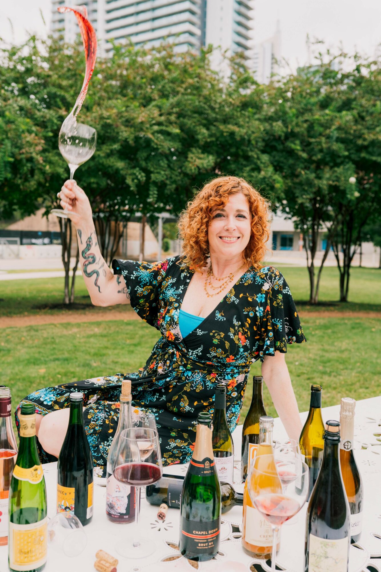Woman with curly hair holding a fork with a glass of wine, smiling outdoors with a table of drinks.