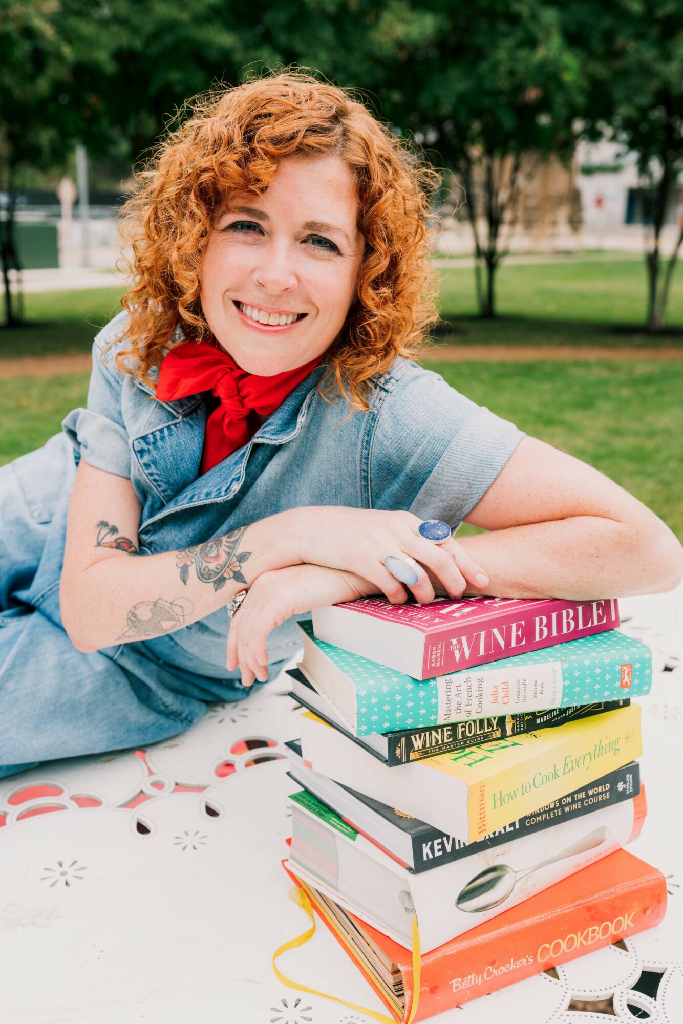 Woman with curly red hair smiling outdoors, sitting at a table with stacked books, wearing a denim shirt and red scarf.