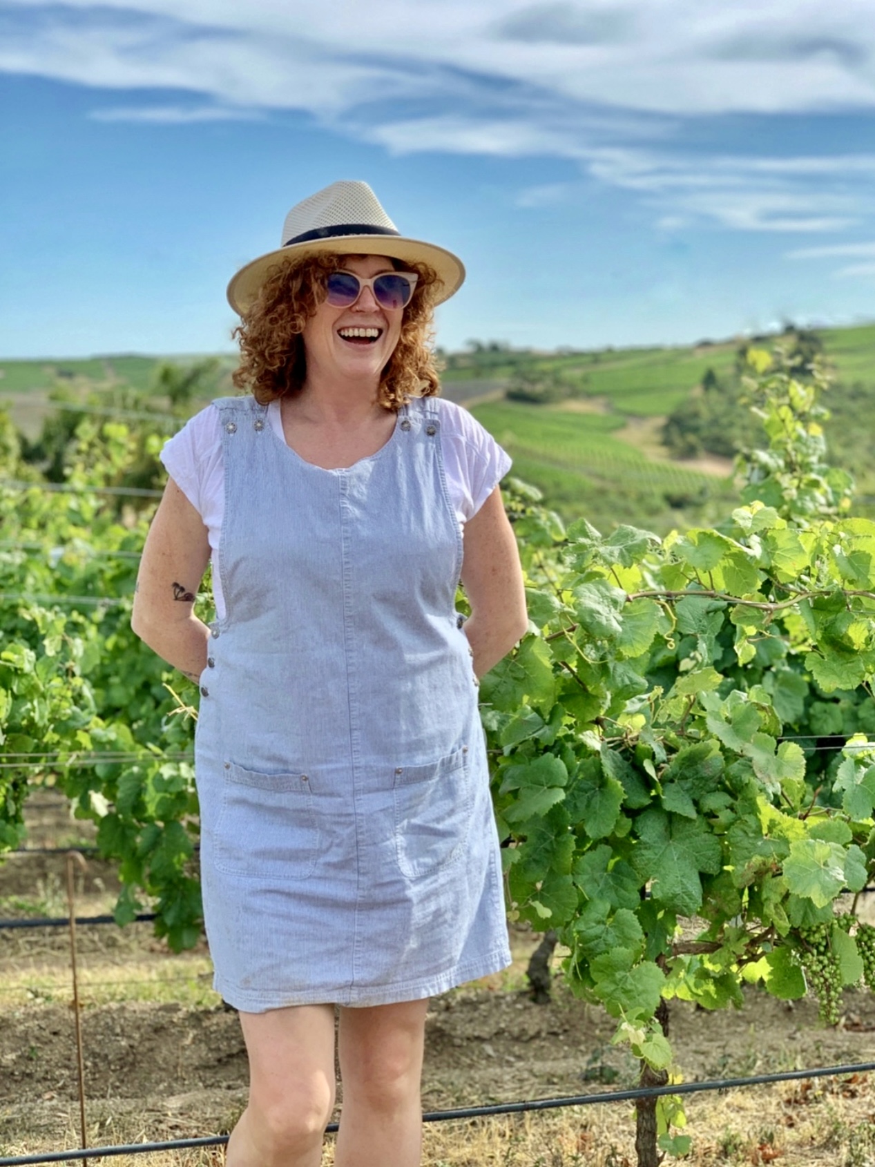 Woman in a light blue dress and hat smiling outdoors in a vineyard with green hills and blue sky.