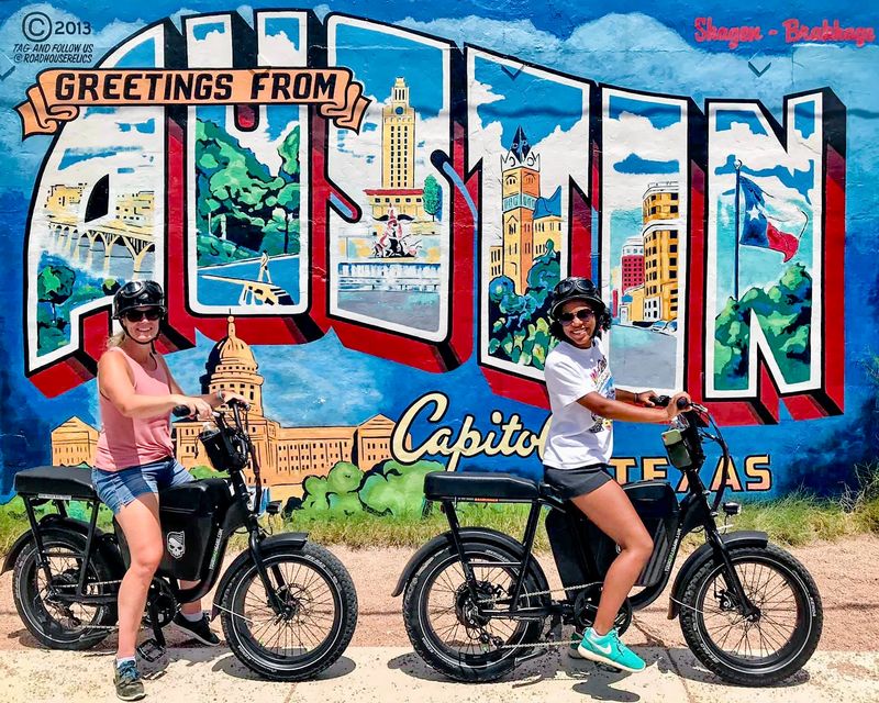 Two women with bicycles in front of a colorful mural of Austin, Texas landmarks, smiling and wearing sunglasses.