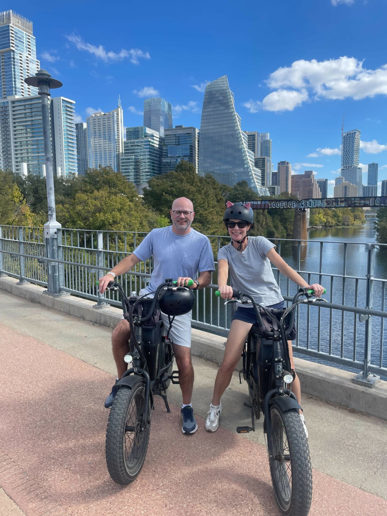 Two people with bicycles on a riverside path with a city skyline in the background, blue sky, and clouds.
