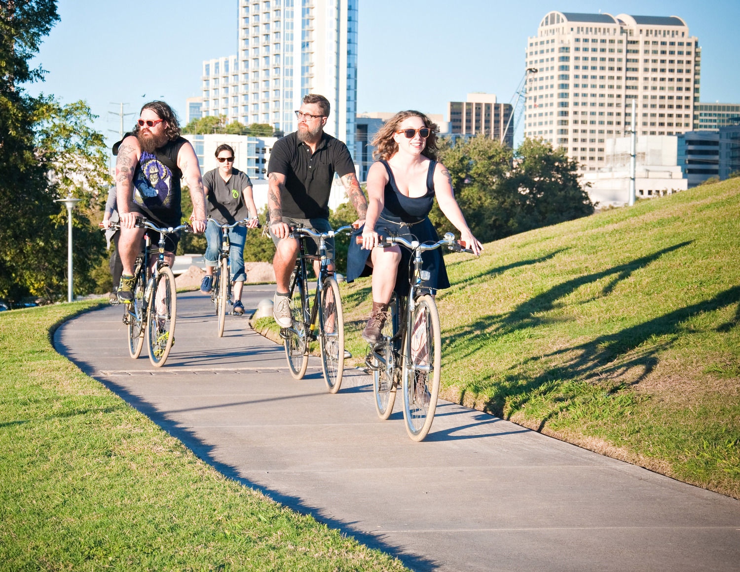 Group of five people riding bicycles on a paved path in a city park with tall buildings in the background.