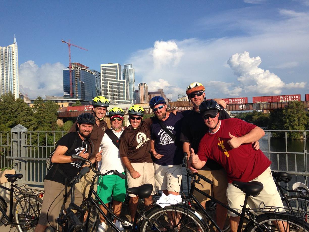 Group of six people with bicycles on a bridge, city skyline in background, clear sky with clouds.