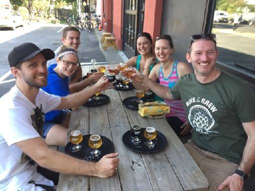 Group of seven people sitting at an outdoor table, raising glasses of beer in a toast, smiling, with food and drinks on the table.