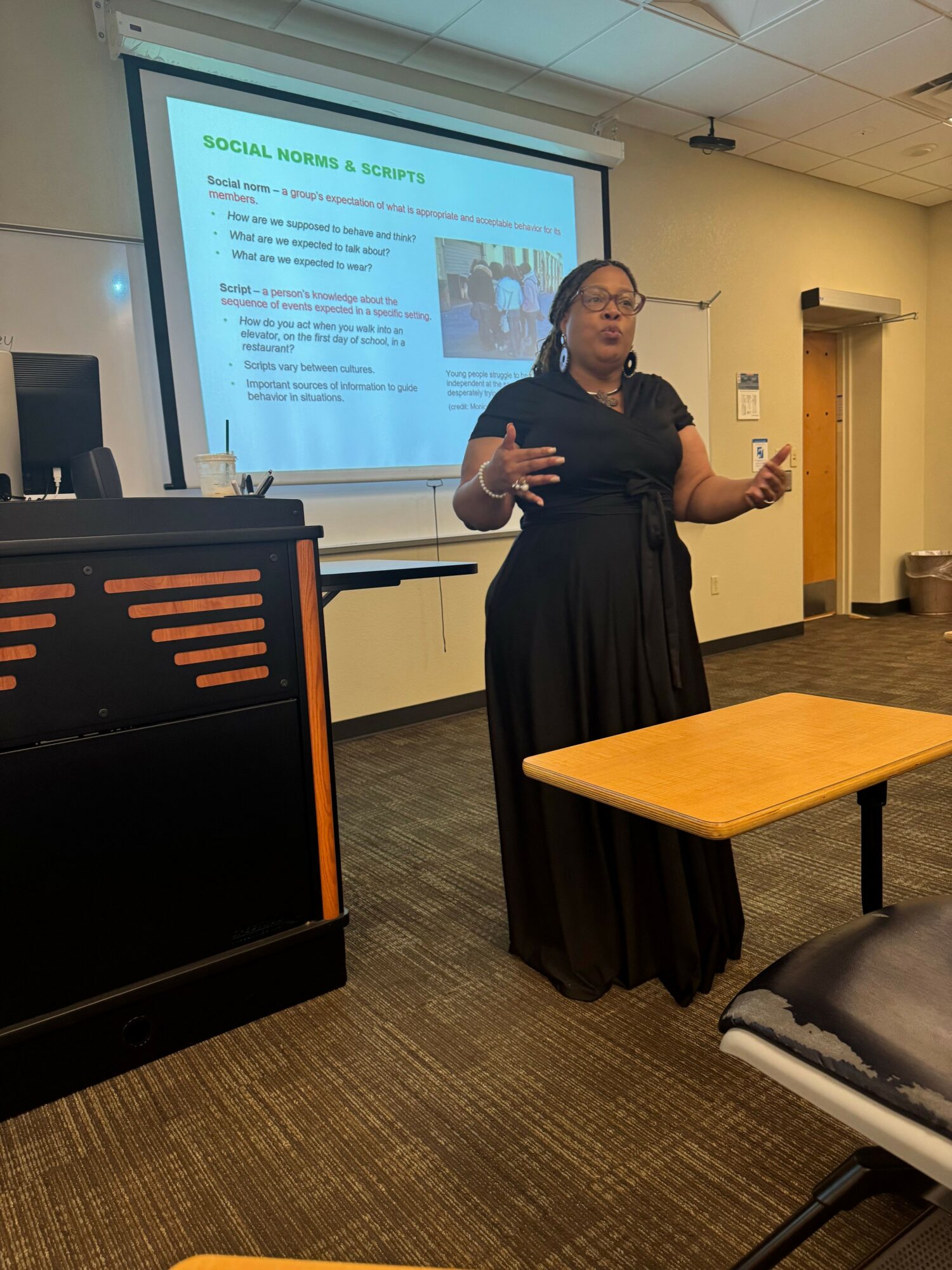 Woman giving a presentation in a classroom, standing near a projector screen with text and images, gesturing with her hands.