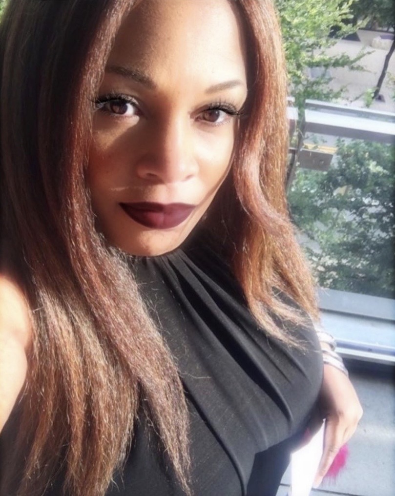Close-up of a woman with long brown hair, dark lipstick, and a black top, standing near a window with trees outside.