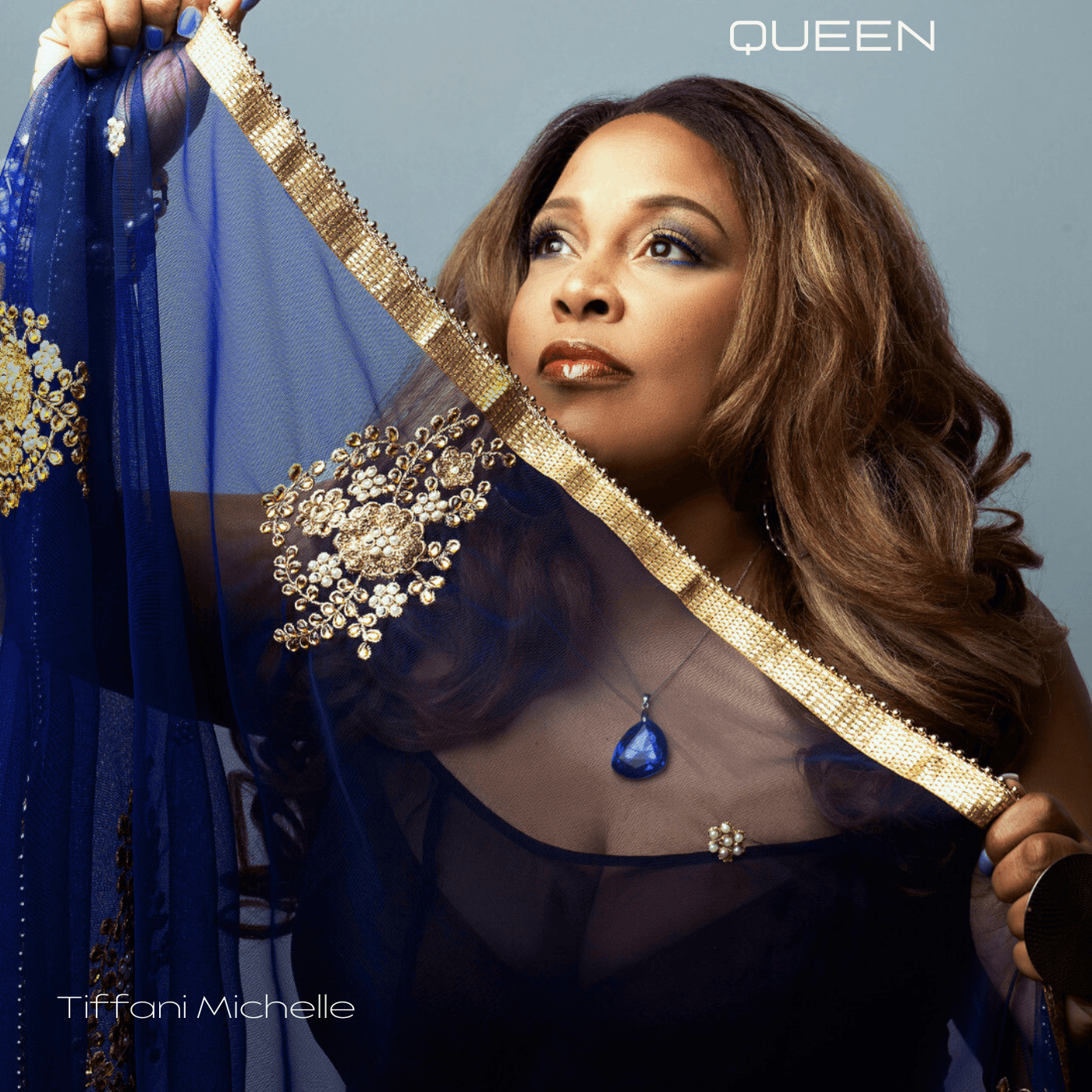 Woman with long wavy hair wearing a black dress and blue jewelry, holding a blue fabric with gold embellishments.