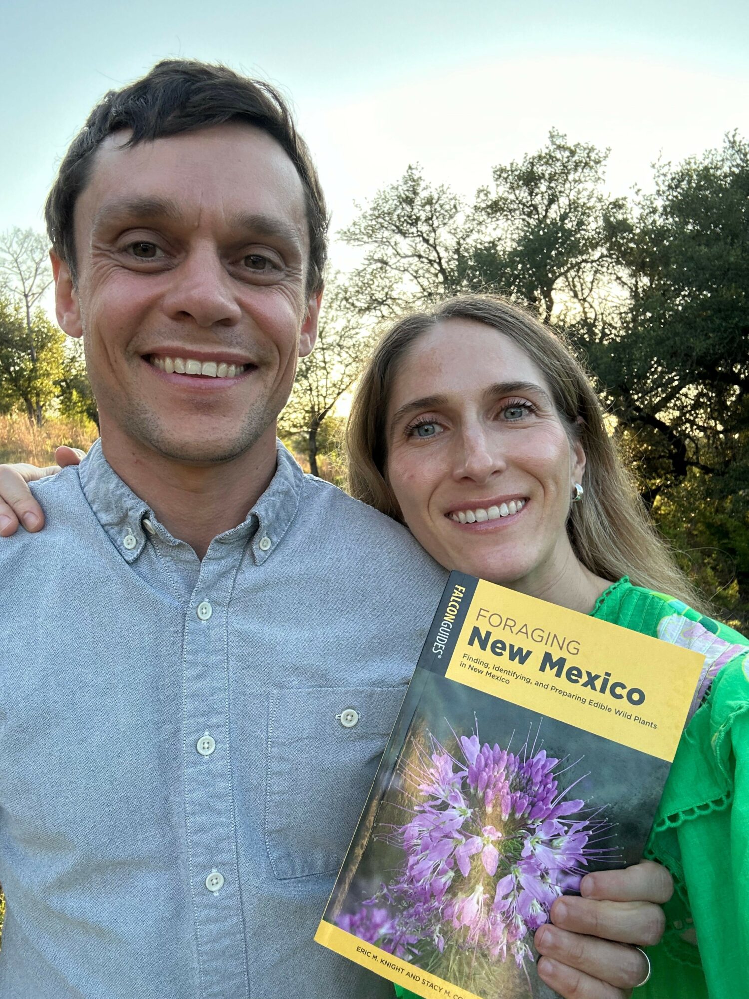 Two smiling people outdoors, one holding a book with purple flowers on the cover, trees and sky in background.