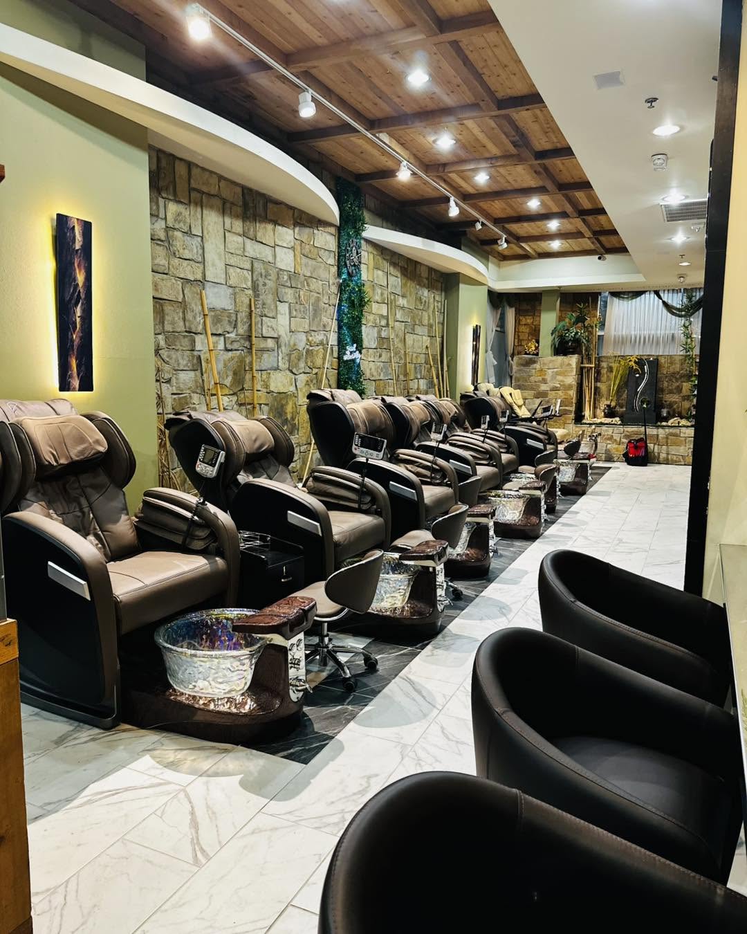 Row of pedicure chairs with foot baths in a salon, wooden ceiling, stone wall, and decorative lighting, viewed from the front.