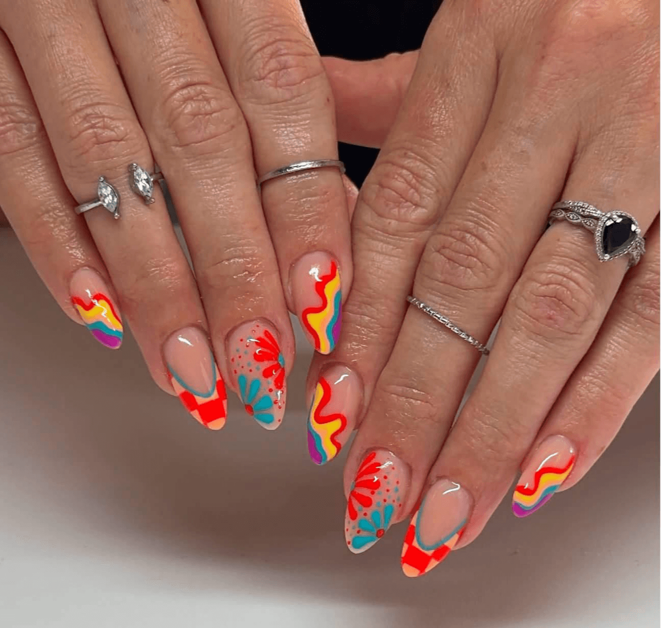 Two hands with decorated nails and rings, displaying colorful rainbow and floral nail art, against a plain background.