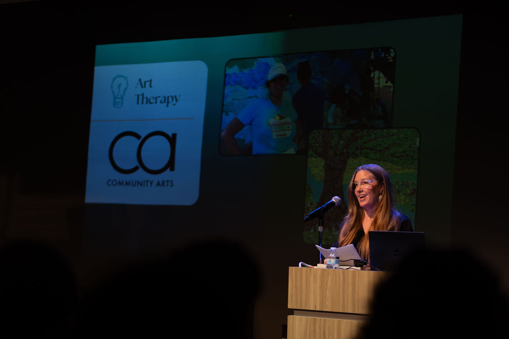 A woman speaks at a podium during a presentation with a screen displaying art therapy and community arts logos.